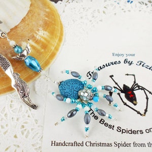 May include: A teal blue and silver beaded spider ornament with a mermaid tail bookmark. The spider has a silver and white beaded body and legs. The bookmark is silver with a mermaid tail design. The text "Enjoy your Treasures by Tiziana The Best Spiders on the WEB Handcrafted Christmas Spider from the Artist" is visible in the background.