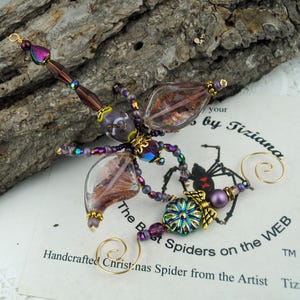 May include: A handcrafted Christmas spider ornament with a dragonfly design. The ornament features iridescent purple, brown, and gold beads, with clear wings and a decorative floral element. The text "The Best Spiders on the WEB" is visible.