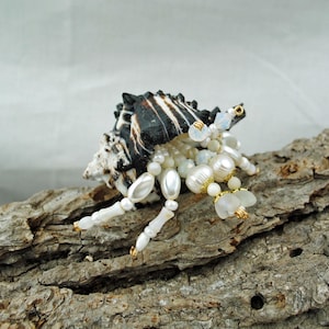 May include: A decorative hermit crab figurine crafted from a dark brown and white striped seashell. The legs and body are constructed from various white and cream-colored beads, including pearls and faceted glass, accented with small gold-toned metal rings.