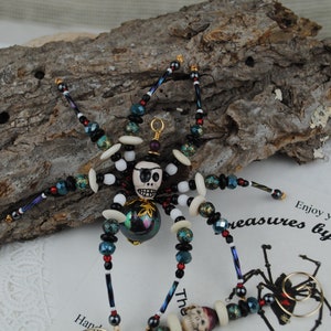 May include: A beaded spider figurine with a skull-shaped head and an eye patch. The spider's body is adorned with black, white, and iridescent beads. The spider is on a piece of wood with a paper that says "Enjoy your treasures by Tiziana."