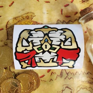 May include: A gold and red sticker of a treasure chest with a keyhole and a red ribbon. The sticker is on a white background.