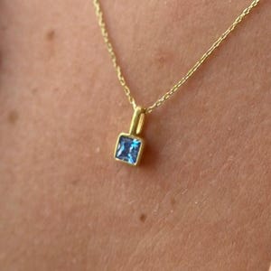 Può includere: Una delicata collana d'oro con un piccolo pendente quadrato con pietra preziosa blu. Il pendente è incastonato in una lunetta d'oro e pende da una sottile catena d'oro. La collana è indossata.