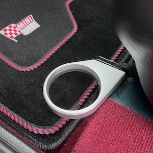 Peut inclure: Un porte-gobelet argenté avec un tapis de voiture noir et rose en arrière-plan. Le tapis porte le texte "MINI COOPER".