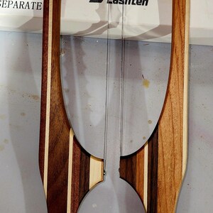 May include: A wooden cheese slicer with a clear wire, featuring a handle with alternating dark and light wood stripes. The word "Lasnten" is visible on the background.