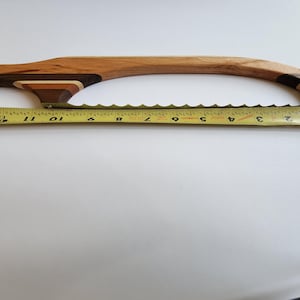 May include: A wooden bread knife with a curved handle and a serrated blade. The handle is made of various wood tones. A yellow tape measure shows the blade is approximately 33 cm long.