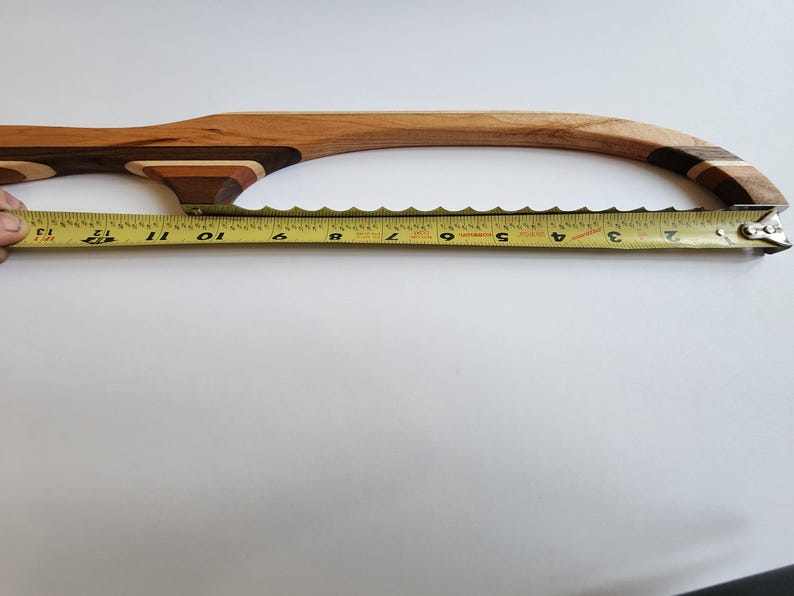 May include: A wooden bread knife with a curved handle and a serrated blade. The handle is made of various wood tones. A yellow tape measure shows the blade is approximately 33 cm long.