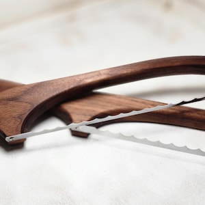 Op de afbeelding: Twee houten broodsnijders met een donkerbruine afwerking. Elke snijder heeft een gebogen handvat en een gekarteld mes van roestvrij staal. De messen staan in een hoek, wat suggereert dat ze zijn ontworpen om efficiënt te snijden.