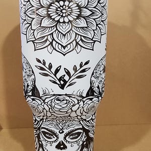 May include: White tumbler with detailed black floral and Day of the Dead designs. The top has a large mandala, the bottom a sugar skull with floral accents. The design is repeated around the tumbler. The tumbler is likely around 18 cm tall.