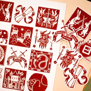 Puede incluir: Impresiones y pegatinas de linograbado rojo y blanco con ilustraciones medievales y fantásticas. Los diseños incluyen conejos con espadas, un caballo, castillos, espadas y un diablo. Algunos diseños incluyen el texto "Audentes Fortuna Iuvat".