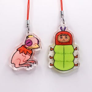 May include: Two acrylic charms with red cords. One charm is a pink bird-like creature with a yellow eye and orange collar. The other is a green caterpillar-like figure wearing a red hood. Both charms have silver-colored metal clasps.