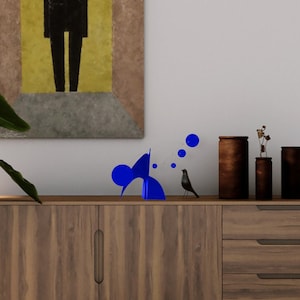 May include: A wooden cabinet with two doors and two drawers. The cabinet is decorated with a blue abstract sculpture and a small black bird. A painting of a black figure in a yellow and brown frame hangs on the wall behind the cabinet.