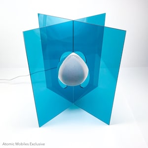 May include: A white speaker sits inside a blue acrylic cube. The cube is made of four panels that are connected at the corners. The speaker is suspended in the center of the cube.