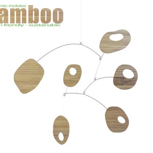 May include: A wooden mobile with five organic-shaped pieces, each with a hole in the center. The mobile is suspended from the ceiling by a thin wire. The pieces are made of light brown wood and have a natural grain pattern. The mobile is labeled "atomic mobiles bamboo earth friendly - sustainable".