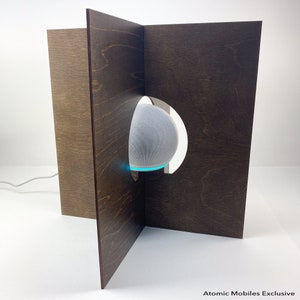 May include: A white smart speaker with a blue light ring is partially visible through a cutout in a dark brown wooden stand. The stand is made of four panels that are joined together at right angles. The stand is on a white surface. Atomic Mobiles Exclusive.