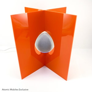 May include: A white smart speaker sits inside a bright orange acrylic stand. The stand is shaped like a cross with four sides. The speaker is in the center of the stand and is visible through a cut-out in the shape of an egg.