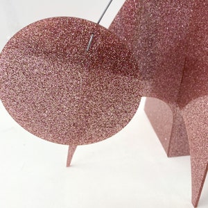 May include: A pink glitter papercraft sculpture with a round, flat, pink glitter circle attached to the top.