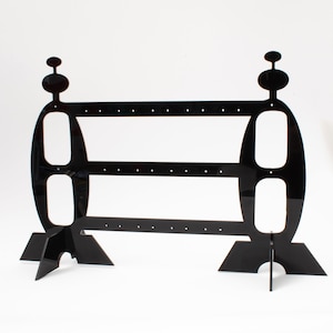 May include: Black acrylic earring stand with two tiers and a unique design. The stand has a rounded base with three legs and a decorative top.