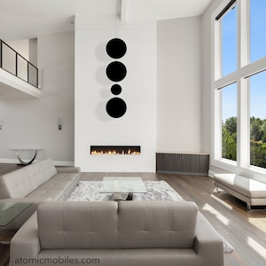 May include: A modern living room with a white fireplace and a black circular mobile hanging above it. The room has large windows with a view of trees and a blue sky. There is a gray couch and a glass coffee table in the room.