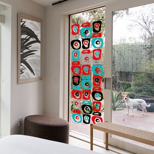 Pu&ograve; includere: Un mobile geometrico colorato fatto di cerchi e quadrati rossi, neri, bianchi e turchesi &egrave; appeso a una finestra. Il mobile &egrave; sospeso dal soffitto ed &egrave; visibile attraverso una porta di vetro. La porta &egrave; aperta e si vede un prato verde e un piccolo cane.