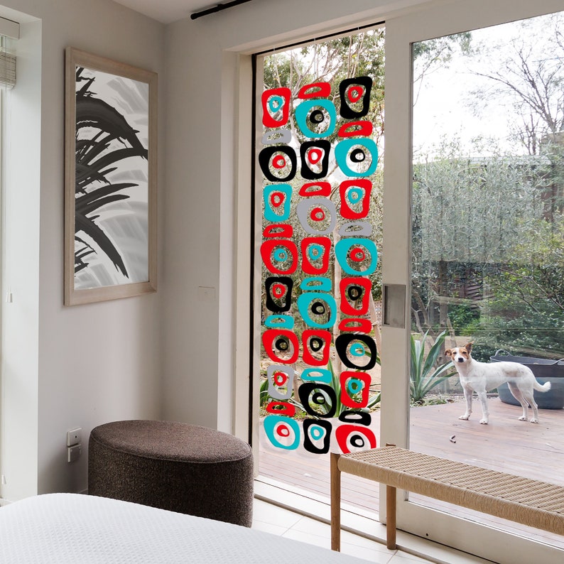 Pu&ograve; includere: Un mobile geometrico colorato fatto di cerchi e quadrati rossi, neri, bianchi e turchesi &egrave; appeso a una finestra. Il mobile &egrave; sospeso dal soffitto ed &egrave; visibile attraverso una porta di vetro. La porta &egrave; aperta e si vede un prato verde e un piccolo cane.