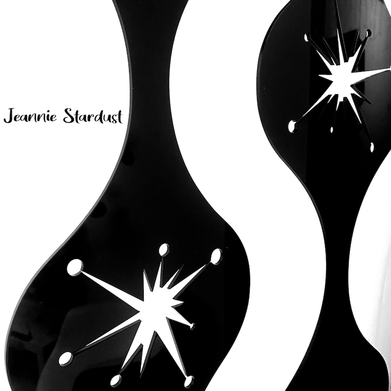 May include: Black mid-century modern decorative wall art with a starburst design. The art features a glossy finish and the text "Jeannie Stardust" in a script font. The design is cut out of the black material, creating a striking contrast.