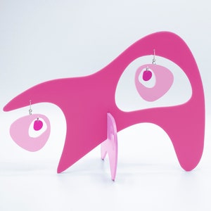May include: Pink and white abstract shaped earrings on a pink display stand. The earrings are made of acrylic and have a modern design.