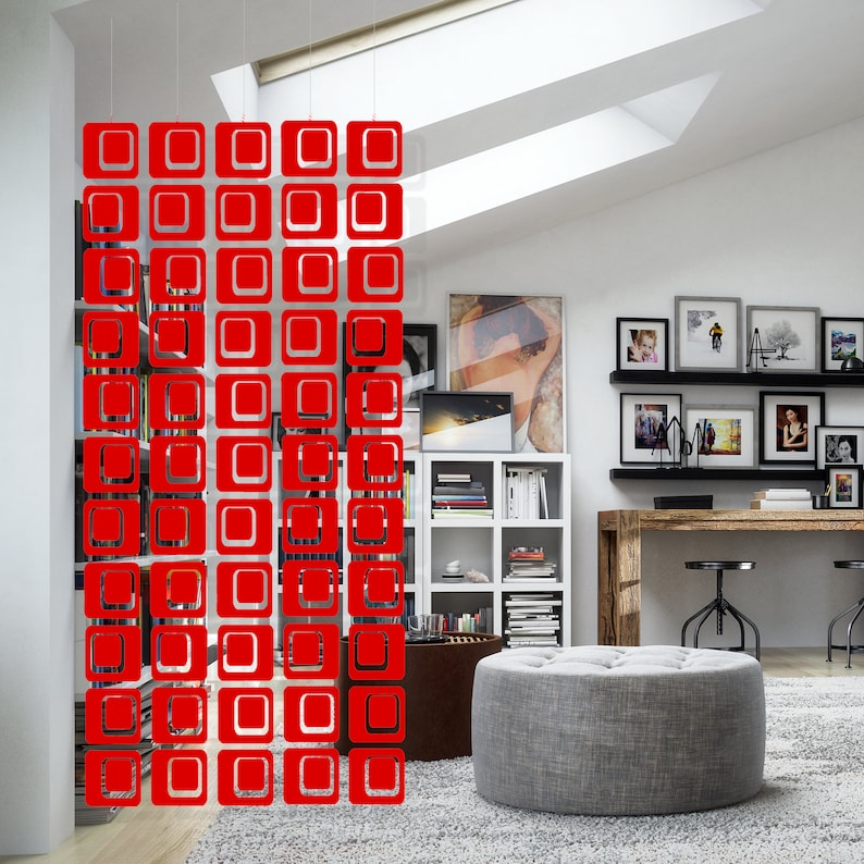 May include: Red room divider made of hanging square shapes. The divider is hanging in a room with a bookshelf, a desk, and a gray ottoman.