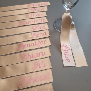 Può includere: Flûte di champagne con nastri personalizzati. I nastri beige presentano nomi come Manuel, Anna e Luise in lettere rosa. Ideali per matrimoni o eventi, questi nastri aggiungono un tocco personale a ogni bicchiere.