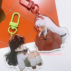 以下が含まれることがあります： アニメのキャラクターが描かれたアクリルキーホルダー2つ。1つは白い髪と赤い衣装のキャラクター、もう1つは黒髪の制服姿のキャラクターです。どちらのキーホルダーにも金属製の留め金が付いています。
