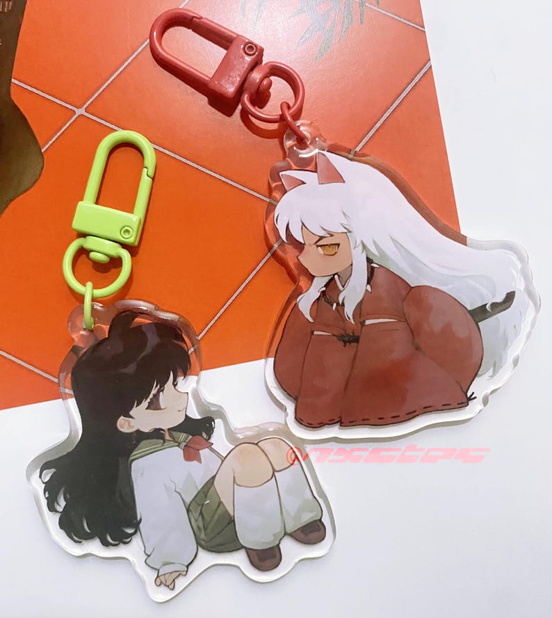 以下が含まれることがあります： アニメのキャラクターが描かれたアクリルキーホルダー2つ。1つは白い髪と赤い衣装のキャラクター、もう1つは黒髪の制服姿のキャラクターです。どちらのキーホルダーにも金属製の留め金が付いています。