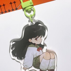 以下が含まれることがあります： 長い黒髪の、白いシャツ、緑のスカート、茶色の靴を履いた、座っているアニメスタイルのキャラクターが特徴の透明なアクリルキーホルダー。キーホルダーには緑色の留め金とリングが付いています。