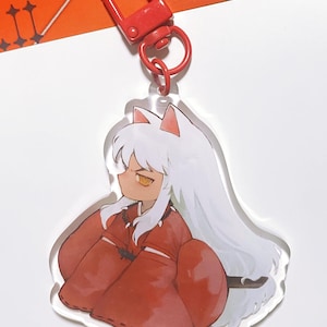 以下が含まれることがあります： 白い髪、赤い服、黄色い目のアニメキャラクターが描かれた、透明なアクリルキーホルダー。キーホルダーには赤い留め金とリングが付いています。キャラクターは座った姿勢です。背景は白とオレンジ色です。