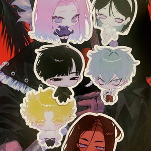 May include: A collection of die-cut stickers featuring anime-style characters with large eyes and various hair colors, including pink, black, yellow, and blue. The stickers are on a dark background, showcasing the detailed artwork and unique designs.