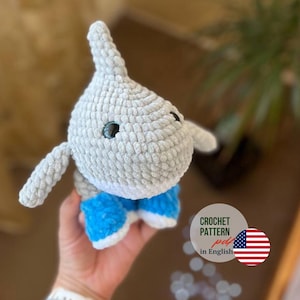 May include: A handmade crochet shark toy in light gray with blue shorts. The shark has small black eyes and a fin. The toy is made of soft, textured yarn. The image includes a graphic that says "CROCHET PATTERN pdf in English".
