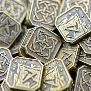 May include: A collection of antique bronze-colored metal coins. The coins have a square shape with rounded corners and feature intricate Celtic knot and mountain designs. The coins are stacked and scattered, creating a textured, close-up view.