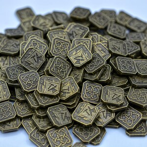 May include: A pile of antique bronze-colored metal coins. Each coin is square with rounded corners and features a stylized design of a mountain, pickaxe, and the letter "T". Some coins have a Celtic knot design.