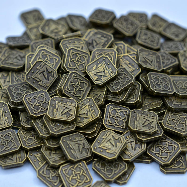 May include: A pile of antique bronze-colored metal coins. Each coin is square with rounded corners and features a stylized design of a mountain, pickaxe, and the letter "T". Some coins have a Celtic knot design.
