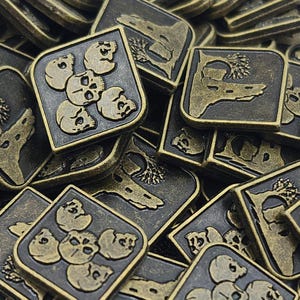 May include: A pile of gold-colored metal tokens with different designs. Some tokens feature a skull and crossbones design, while others have a tree and a rock formation. The tokens are square shaped and have a black background.
