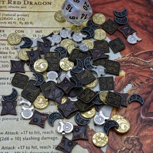 May include: A selection of metal gaming tokens and a clear, multi-sided die. The tokens, in silver, gold, and bronze, display designs like dragons, crests, and crescent moons. The items are scattered on a surface with text.