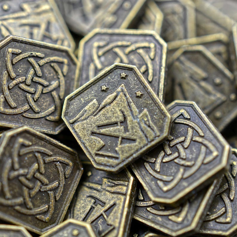 May include: Close-up of a pile of antique bronze-colored metal coins. The coins have a hexagonal shape with intricate Celtic knot and mountain designs. The coins are likely for a board game or collectible.