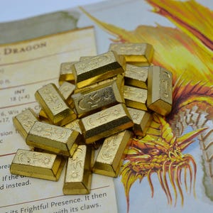 May include: A collection of gold-coloured, rectangular game pieces with engraved symbols, including moons and suns. The pieces are scattered on a game board with text and a dragon illustration. These are likely for a tabletop role-playing game.
