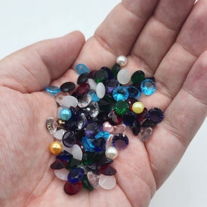 May include: A variety of colorful glass rhinestones in various shapes and sizes, including round, square, and oval. The rhinestones are scattered on a hand, creating a vibrant and sparkly display.