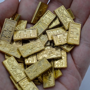 May include: A close-up of numerous small, gold-coloured rectangular objects, possibly dominoes, with detailed embossed patterns. The pieces are held in a hand, showcasing their intricate designs. The objects appear to be game pieces or decorative items.