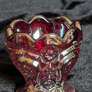 May include: A small, red glass dish with a scalloped rim and a decorative pattern. The dish has a iridescent finish and is perfect for holding small items.