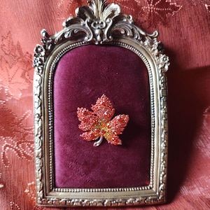 May include: A vintage-style, ornate gold-colored frame with a burgundy velvet backing. A sparkling, red and yellow jeweled maple leaf brooch is pinned to the velvet. The frame has an arched top and is set against a patterned, reddish-orange fabric.