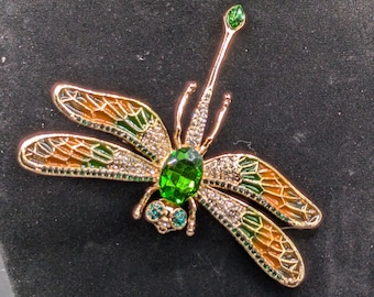 May include: A gold dragonfly brooch with green, orange, and clear crystal detailing. The brooch features a large green central stone and smaller green stones for eyes. The wings are decorated with orange and green enamel.