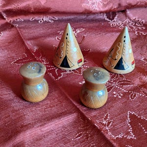 May include: A set of four wooden salt and pepper shakers. Two are cylindrical with carved diamond patterns, and two are cone-shaped with painted teepee designs. The shakers are on a red patterned surface.