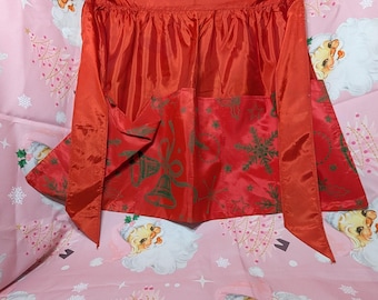 May include: A red Christmas apron with green snowflake and bell designs. The apron has a gathered waist and long ties. The apron is made of a shiny, possibly satin-like material.