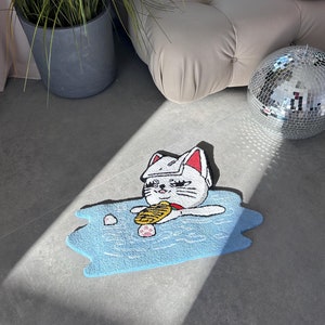May include: A blue bath mat featuring a white cat wearing a white hat and holding a yellow fish. The cat is in a blue bath with white bubbles. The mat is on a grey floor.