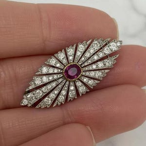 May include: An elongated, marquise-shaped brooch featuring a central oval-cut deep red ruby surrounded by a halo of small, round, clear white diamonds set in a radiating sunburst pattern of silver-toned metal with a fine milgrain edge detail.